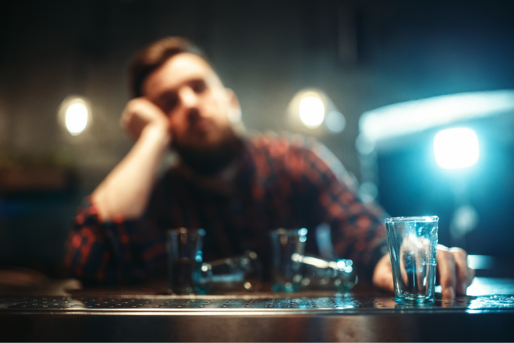 Close up of a shot glass with a blurry drunk man sitting at a bar in the background