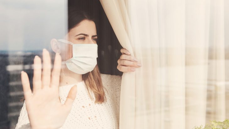 Woman wearing a mask in isolation seen from a glass window.