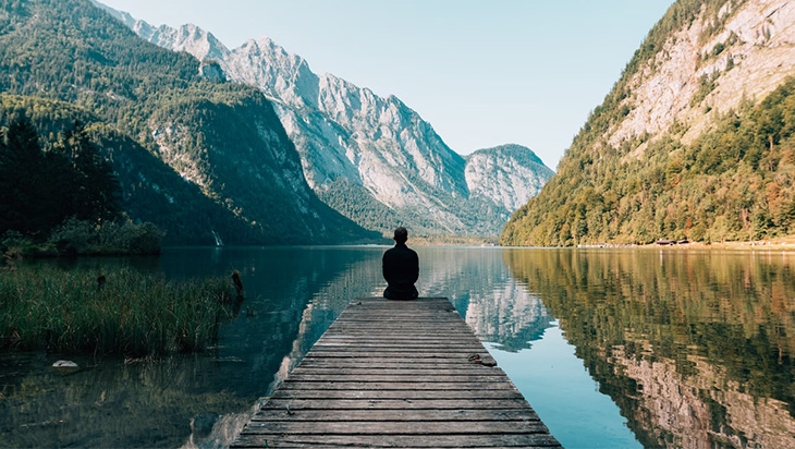 A man meditating on the edge of a wooden dock as he looks at a picturesque lake.