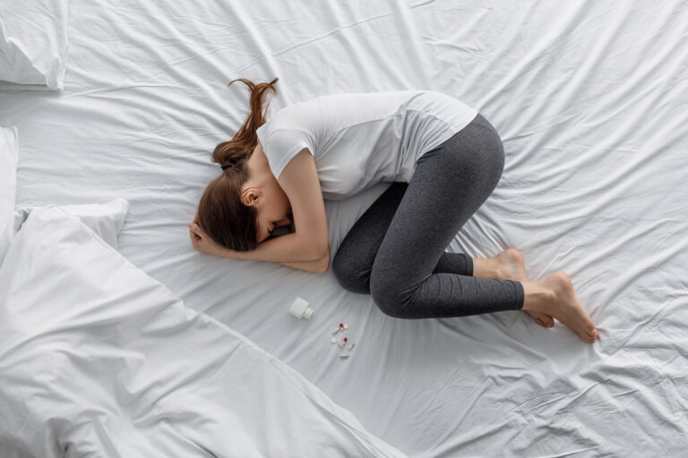 A woman curled up on a bed with a pill bottle and tablets beside her