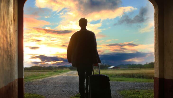 A man holding a suitcase walks through an archway into an open field during sunset