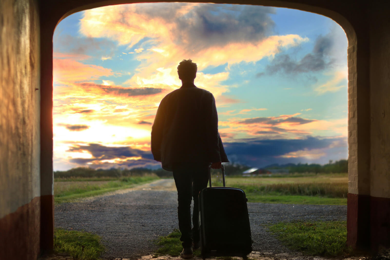 A man holding a suitcase walks through an archway into an open field during sunset