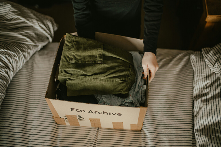 A person placing a cardboard box full of folded clothes on a bed with striped sheets