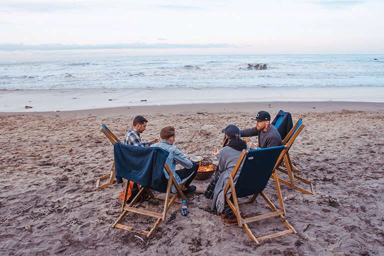 A group of four adults sitting in chair chairs around a fire on a beach