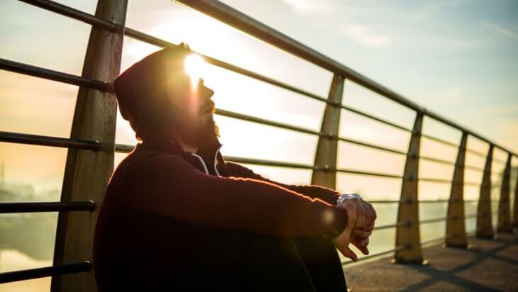 A hooded man sitting on a bridge leaning against the railing while the sun rises in the background