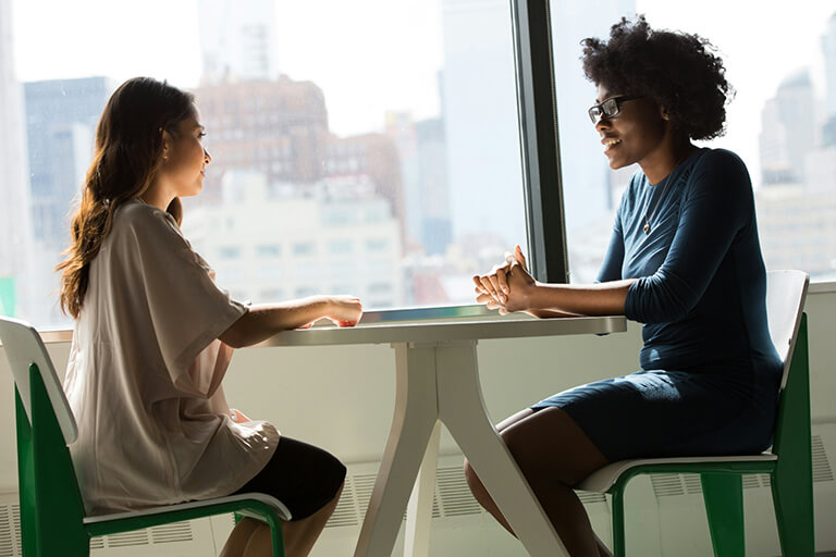 Two women talking at a table by a large window overlooking the city