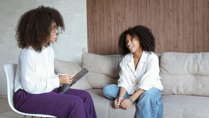 A female therapist sits across from a client during a therapy session, both smiling.