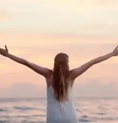 A woman with outstretched arms facing the ocean at sunset