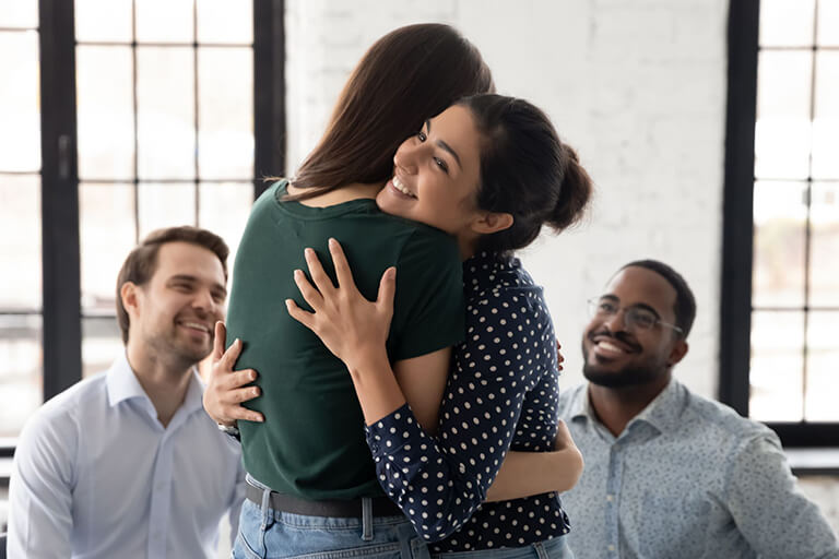 Two people embrace, while two others watch supportively during a group counselling session