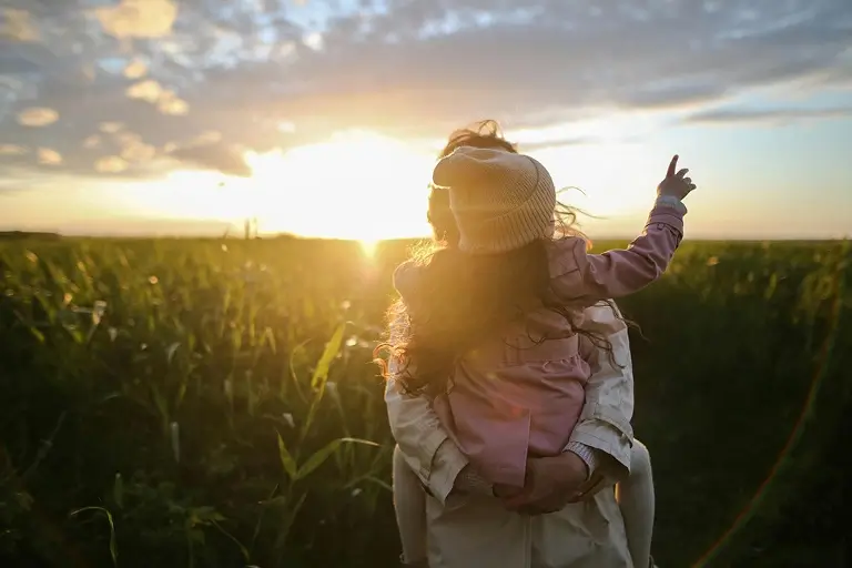 Parent holding child while watching sunset over a field.