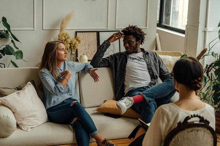 A man and a woman sit on a couch opposite a therapist in a couples therapy session.