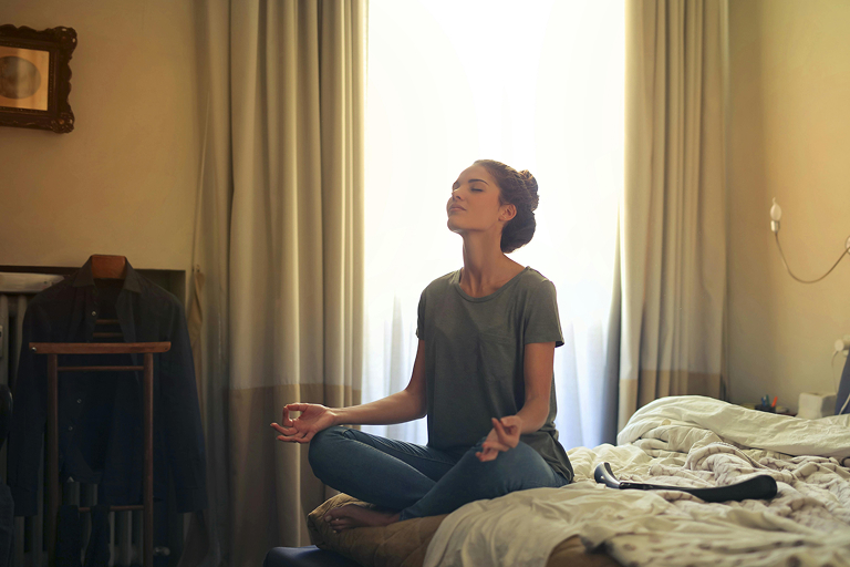 A woman meditates on a bed with her legs crossed and eyes closed.